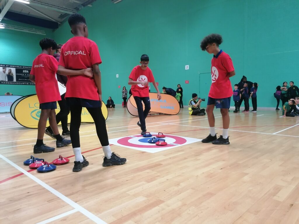 A group of young leaders in a sports hall wearing red T-Shirts and black tracksuits/shorts. They are officiating a new age Kurling match, with the stones in a target area and children watching in the background.