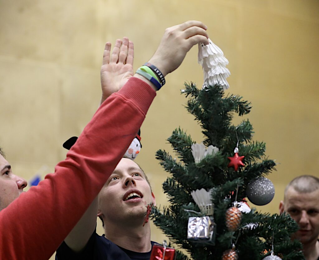 Two people look at a Christmas tree whilst placing a badminton shuttlecock at the top.