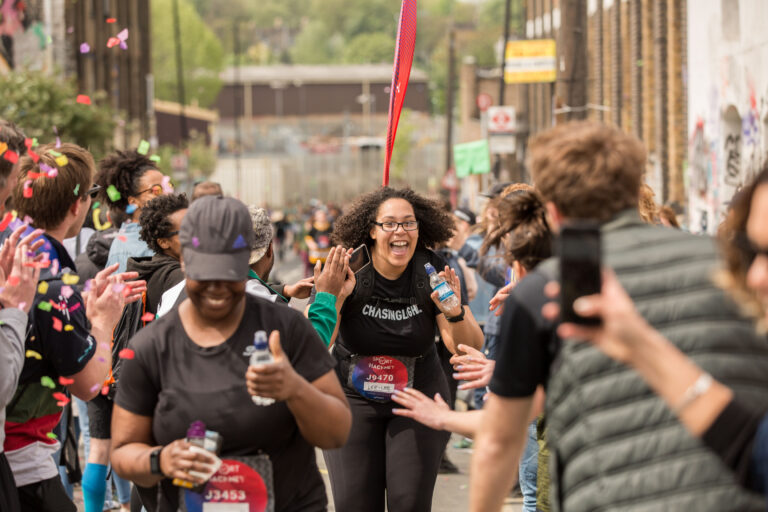 A woman smiles at the camera whilst running a half-marathon. They are wearing all black clothing.